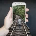 A person holding up a smartphone which has a man walking along train tracks emerging out of it.